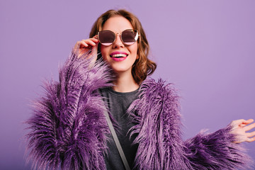 Magnificent female model in amazing purple fur coat looking to camera through sunglasses. Laughing excited caucasian girl with shiny hair posing in trendy winter outfit.