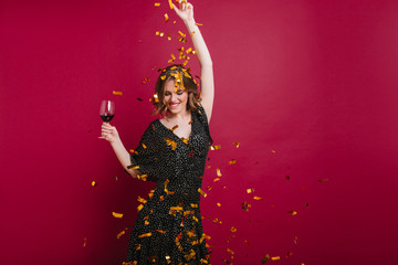 Joyful attractive female model dancing with pleasure and throwing out golden confetti. Adorable caucasian girl in black clothes having fun at festive and tasting red wine.