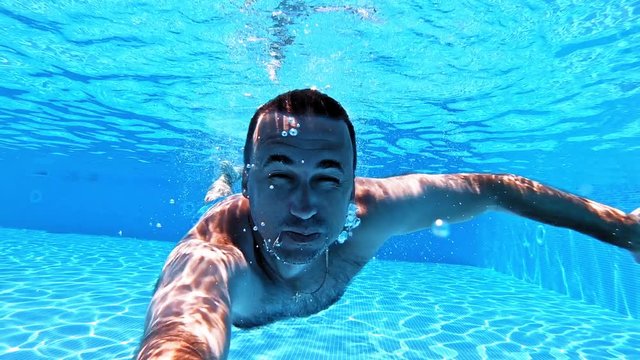 Man swims with underwater camera inside the swimming pool. Transparent water background of a pool and male adult swimming underwater and looking into camera.