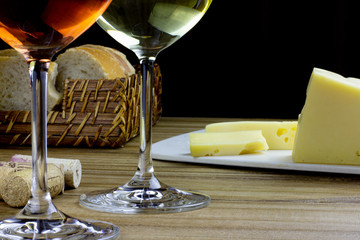 White wine glass and rose wine glass on wooden table with basket of bread, cheese and corks on black background