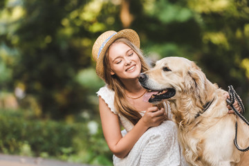 beautiful girl in white dress and straw sitting near golden retriever and smiling while looking at dog