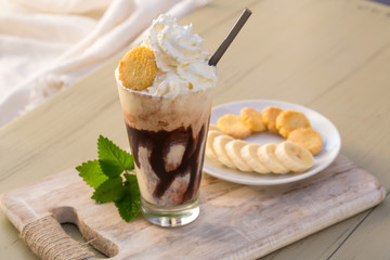 Tasty milkshake drink in glass mug with plastic tube and slice of banana on chopping board on wood table