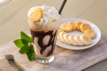 Tasty milkshake drink in glass mug with plastic tube and slice of banana on chopping board on wood table
