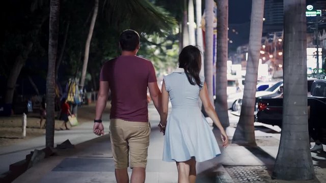 Romantic and love relationships. Young couple male and female walking along the promenade at night street in Pattaya, Thailand, rear view.