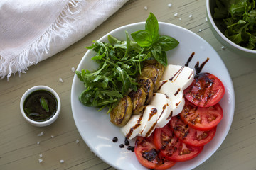 Caprese salad with mozzarella, tomato, basil and grilled eggplant on white plate. Top view on background