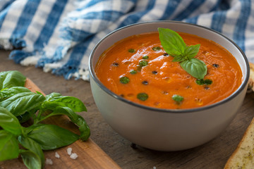 A bowl of fresh tomato soup in white ceramic bowl, garnished with basil, croutons, seasoning and a drizzle of olive oil, and served with crusty bread.