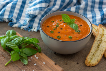 A bowl of fresh tomato soup in white ceramic bowl, garnished with basil, croutons, seasoning and a drizzle of olive oil, and served with crusty bread.