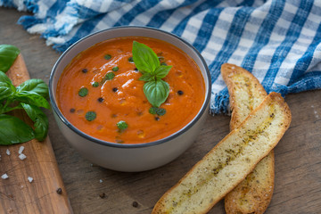 A bowl of fresh tomato soup in white ceramic bowl, garnished with basil, croutons, seasoning and a drizzle of olive oil, and served with crusty bread.