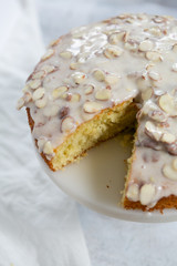 slice of almond olive oil cake with brown butter icing and sliced almonds, dripping icing on cake, marble and wood cake stand, styled bakery