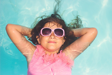 Cute little girl with pink sunglasses, smiling and laying relaxed in the pool; summertime background with copy space