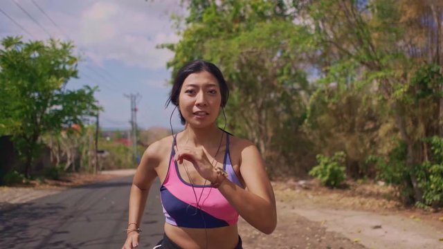 Slow motion frontal pull back gimbal shot in dolly style of young exotic fit and beautiful Asian Indonesian woman running on asphalt road doing jogging workout in sport and healthy lifestyle concept