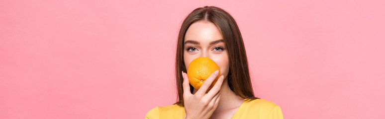 panoramic shot of young woman eating orange and looking at camera isolated on pink