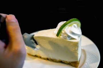 Image of hand holding a teaspoon is dividing a cake from a lime cream cake on a white ceramic plate and black background.