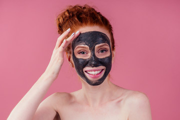 redhaired ginger teenager girl with black clay mask on her pretty face on pink studio background