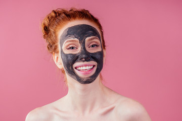 redhaired ginger teenager girl with black clay mask on her pretty face on pink studio background