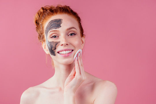 redhaired ginger teenager girl remove with cotton black clay mask on her pretty face on pink studio background