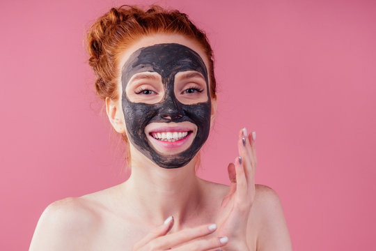 redhaired ginger teenager girl with black clay mask on her pretty face on pink studio background