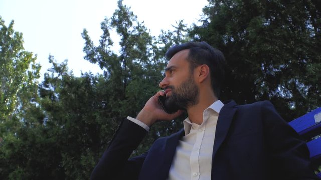 businessman using a mobile phone in a park on a bench. man talking on a cell phone. bearded man talking on a smartphone. young attractive bearded man is sitting and talking on a cell phone with a frie