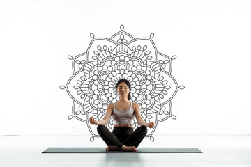 calm thai woman practicing yoga on yoga mat near mandala ornament on white