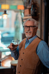 Grey-haired man wearing brown vest drinking whisky