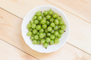 Green berries of gooseberry in white bowl in center on wooden background with copy space.