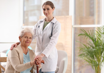 Nurse assisting elderly woman with cane indoors. Space for text