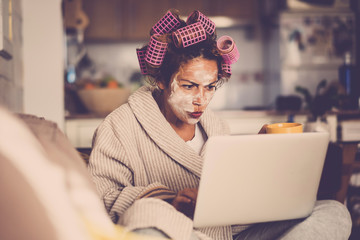 Angry and beautiful young people middle age caucasian woman with wellness cream mask on the face - looking the web for home made remedy - curlers for beauty make up work
