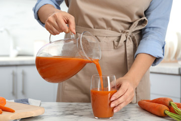 Woman pouring tasty carrot juice from jug into glass at table indoors, closeup