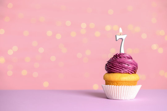Birthday cupcake with number seven candle on table against festive lights, space for text