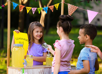 Little girl selling natural lemonade to kids in park. Summer refreshing drink - obrazy, fototapety, plakaty