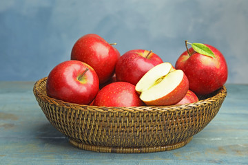 Wicker bowl with ripe juicy red apples on wooden table against blue background