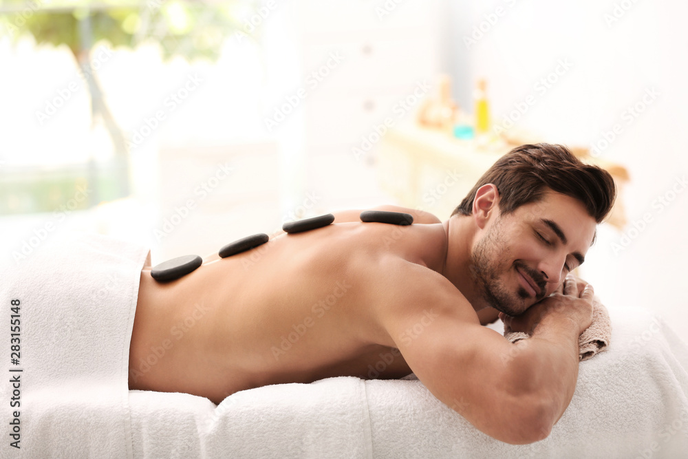 Wall mural Handsome young man receiving hot stone massage in spa salon, space for text