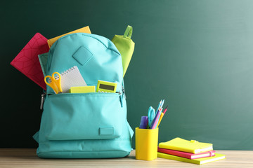 Backpack full of different school stationery on table near blank chalkboard. Space for text