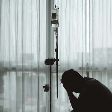 Silhouette Stressful And Sad Asian Man Sitting And Worried While Having Medical Treatment With Hospital Curtain And Window As Background.