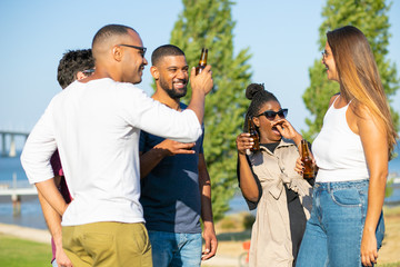 Joyful best friends laughing while drinking beer in park. Five smiling young people relaxing during picnic. Leisure concept