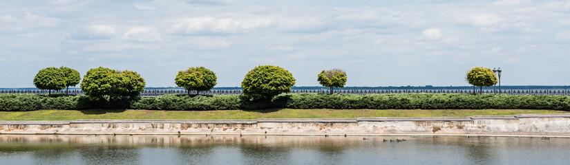 Fototapeta na wymiar panoramic shot of green leaves on trees near lake against sky with clouds in summer