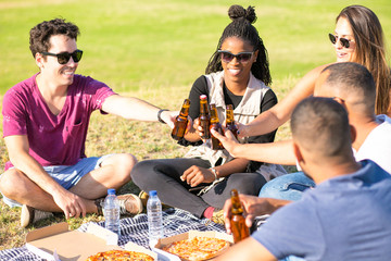 Smiling young people clinking beer bottles in park. Happy friends relaxing on meadow and drinking beer. Leisure concept