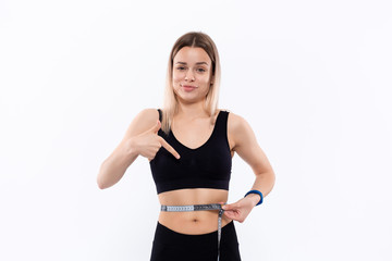 Obraz premium Young blond woman in a black sportswear with smart watches is happy pointing at the tape measure standing over white background. Checking waist after workout.