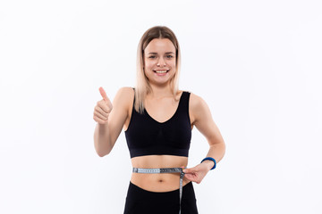 Fototapeta na wymiar Young blond woman in a black sportswear with smart watches is happy checking waist after workout with the tape measure standing over white background.
