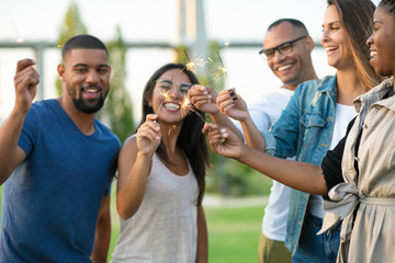 Cheerful young friends holding sparklers. Happy young multiethnic men and women gathering together and celebrating in park. Party concept