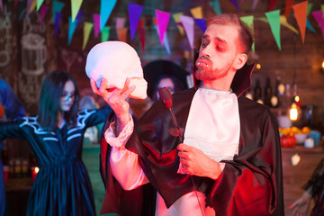 Attractive young man dressed up like dracula holding a black rose looking at his human skull at...