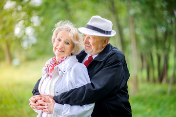 Fototapeta premium happy elderly couple hugging tightly while standing in a park at sunset, smiling and looking at the camera
