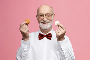 Which one do you like? Isolated image of cheerful energetic senior retired male wearing glasses and bow tie, smiling broadly, holding colorful macarons in each hand, offering you to have some