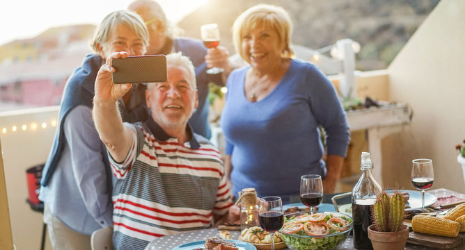 Happy seniors friends taking a selfie photo with smartphone camera at barbecue dinner in house terrace - Mature people having fun with new trend technology - Focus on mobile cell phone