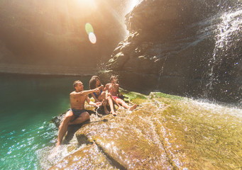 Group of friends having fun under waterfalls river - Young people swimming inside emerald water...