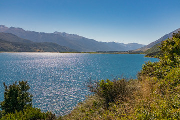 The sparkling waters of a pristine lake in Otago, New Zealand, surrounded by jagged mountain peaks against a clear blue sky and remote bushland