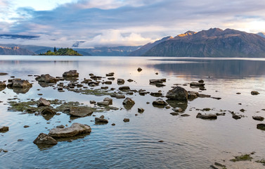 A low-level view of Lake Wanaka early in the morning as the sun rises on the hills in the distance, taken from the water's edge, rocks in the foreground.