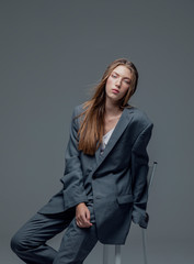 Portrait of weary young beautiful woman in oversized man's suit sitting isolated grey background