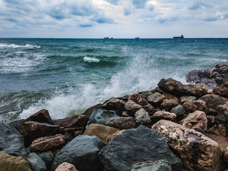 Sea waves with foam beating large stones on the shore on a summer day with ships on the horizon and cloudy sky.