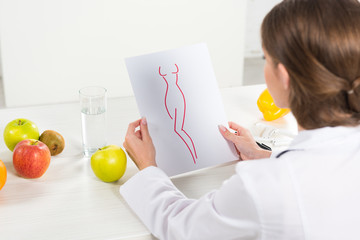 cropped view of dietitian in white coat holding paper with perfect body image at workplace